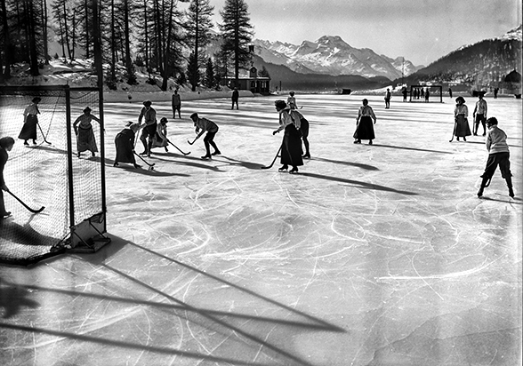 Das schwarz-weiss Bild zeigt Männer und Frauen auf einem grossen Natur-Eisfeld, einem zugefrorenen See. Die Personen tragen Schlittschuhe und halten Stöcke. Sie spielen Bandy, eine frühe Form von Eishockey mit einem Ball. Im Hintergund sieht man eine Alpenkette und links im Hintergrund an das Eisfeld angrenzend einen kleine Wald.