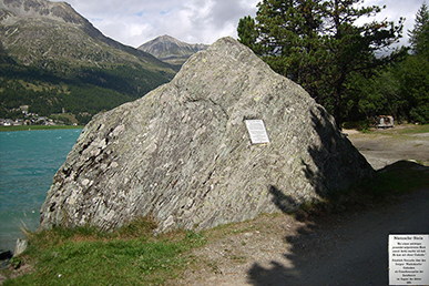 Das Bild zeigt einen pyramidförmigen Felsblock ab dem auf einer Platte eine Inschrift angebracht ist. Im Hintergrund sieht man die Landschaft von Silva Planta mit See, Tanne und Bergen.