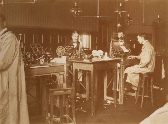 Das Bild zeigt drei Herren die im Berner Telgrafenamt im Jahr 1900 arbeiten. Auf einem erhöhten Holztisch stehen die Telegrafenaparaten, um die gruppiert die drei Männer auf erhöhten Hockern sitzen.