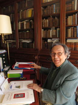 La photographie montre Jean Starobinski assis à sa table de travail, au milieu de sa bibliothèque, avec des livres ouverts devant lui.