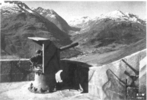 « Canon de 8,4 cm sur affût à pivot central (dit «à crinoline») dans le voisinage immédiat du fort Bäzberg. En arrière, le village de Hospenthal et la route du Saint-Gotthard », photographe inconnu, Walter Lüem, « Les origines de la fortification du Saint-Gothard : "Qui tient le Saint-Gothard, tient la Suisse" », in : Revue Militaire Suisse 137 (1992), fasc. 5, pp. 35-41, © Revue militaire suisse © Revue militaire suisse, photo : p. 38