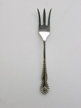 The photograph shows a small, artistically decorated, three-pronged silver fork. The left and right prongs are both curved slightly outwards. The fork shaft is somewhat broader towards the end and has ornament-like decorations.