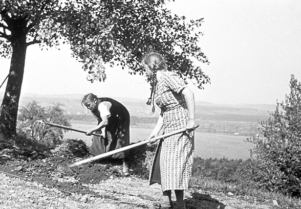 The black and white photo shows two women working in the fields, with a lake and open countryside in the background. The older woman on the left of the photo is bending forward and vigorously hoeing. The woman on the right appears to have just completed her movement and is standing up straight holding a hoe. Both women are fully focused on the task at hand.