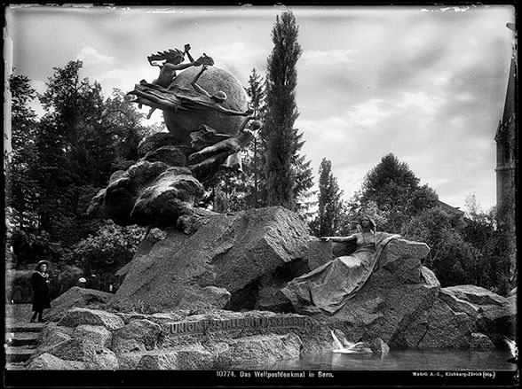 The picture shows the stony monument of the Universal Postal Union on the “Kleine Schanze” in Bern. In the upper left corner you can see the symbol of the Universal Postal Union, which shows different people transmitting letter mail around the globe. On the right side of the picture is a stone figure, which benevolently looks at the hustle and bustle in the upper right corner of the picture. In front of the stone monument a small pond can be seen.