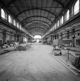 The hall of Zurich’s Herdern slaughterhouse resembles a cathedral. This photograph shows the central nave. The hall has a barrel-vaulted ceiling. Its clerestory is punctuated by arched lattice windows which make the space light and elegant. Construction machinery, a skip and other building tools stand around on the floor. The photograph dates from the years 1985–87, during which the hall was renovated and transformed.
