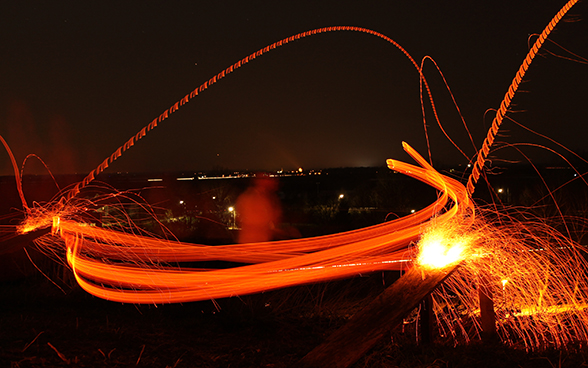 The image shows trails of light in the night sky produced by a young man who starts by swinging a glowing wooden disc on a long rod before hurling the disc into the valley below using a catapult.