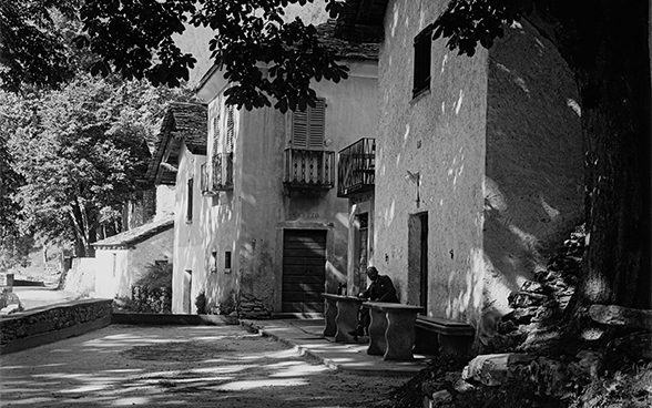 À gauche de l’image, un large chemin ombragé passe devant plusieurs maisons en pierre accolées les unes aux autres, qui abritent des grotti. Devant la maison, un homme dont on devine à peine les traits est assis à une table en pierre. Au bord de la route, à droite, se trouve un châtaignier dont une branche s’étend au-dessus de la route.