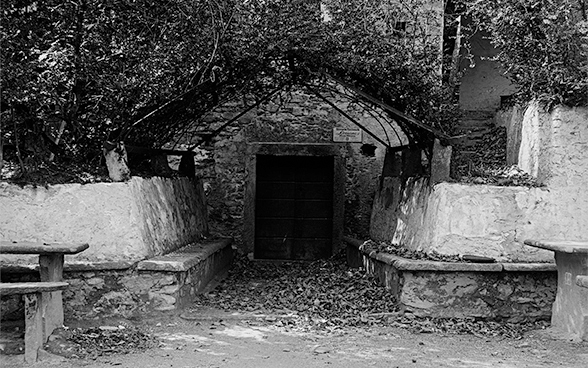 Vue de face d’un grotto de deux étages, entouré d’arbres dont les branches sont encore nues. La porte d’entrée du grotto est fermée, des feuilles mortes couvrent le sol. À gauche et à droite de l’entrée, un muret de pierre qui invite à s’asseoir.