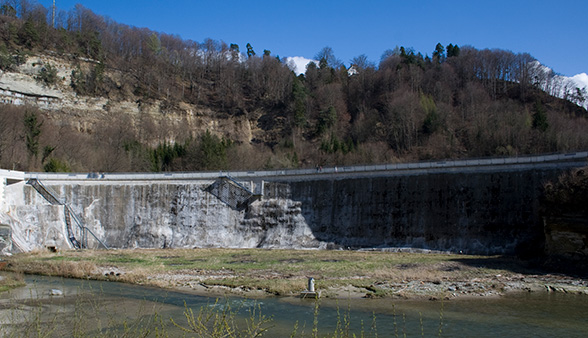 La photo en couleur montre le barrage de la Maigrauge, haut de 24 mètres et légèrement incurvé, vu d'en bas. On voit à l'arrière-plan un ciel bleu et l'imposante paroi rocheuse, en partie boisée, surplombant la Sarine. La photo a été prise en hiver car les arbres n'ont pas de feuilles.