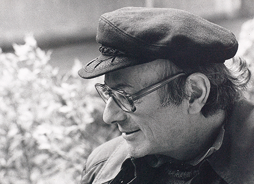 Walter Vogt con cappellino a visiera sorride mentre guarda in basso a sinistra  - sullo sfondo un cespuglio in fiore.