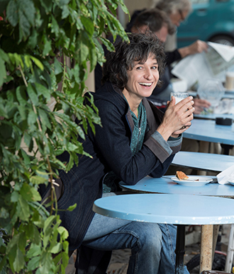 Nadja Sieger sitzt in Gartenrestaurant mit Glas in der Hand, lachend.